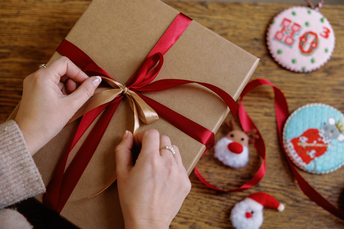 Person tying a red ribbon on a brown gift box with Christmas decorations in the background.