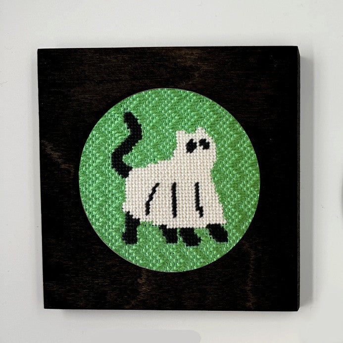 A needlepoint ornament of a cat dressed up as a ghost, displayed in a black frame