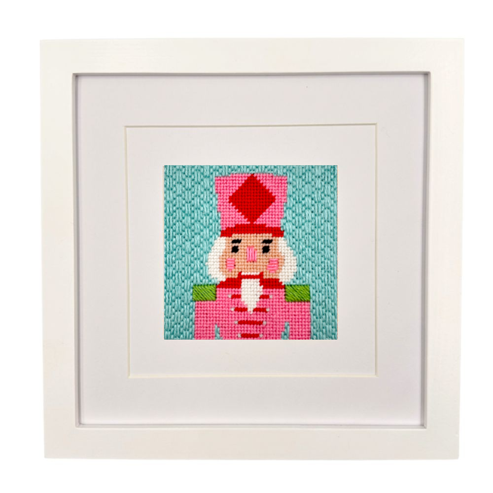 A framed needlepoint canvas featuring a nutcracker character on a teal background, presented in a white square frame with a white mat.