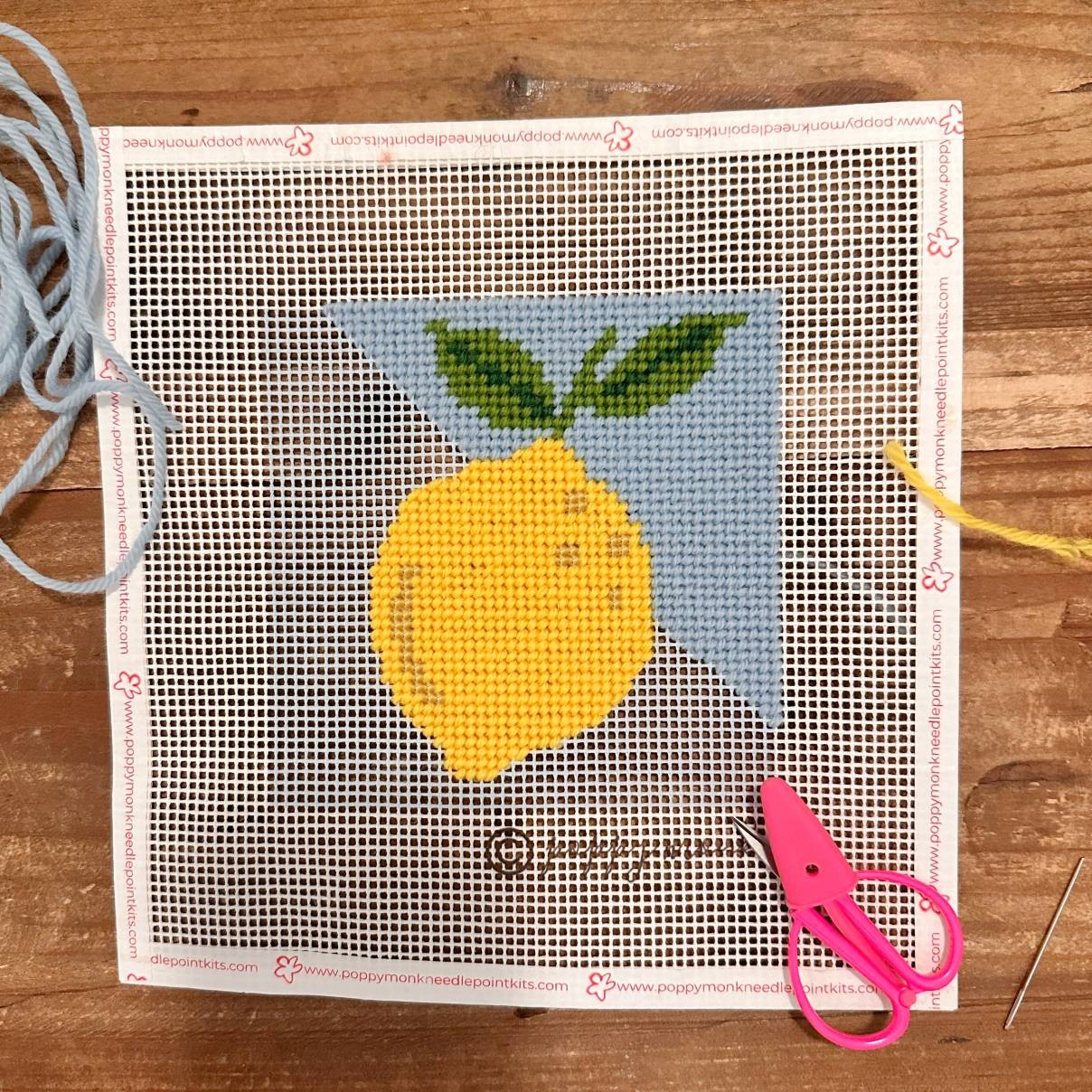 Needlepoint project with a lemon design on a wooden surface with scissors and thread.