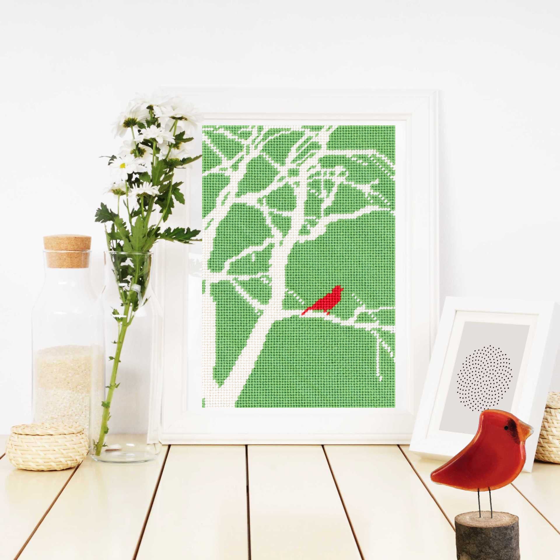 The Red Bird design in a white frame leaning against the wall, styled on a light wood tabletop with a glass bottle, white flowers, and a small red bird figurine.