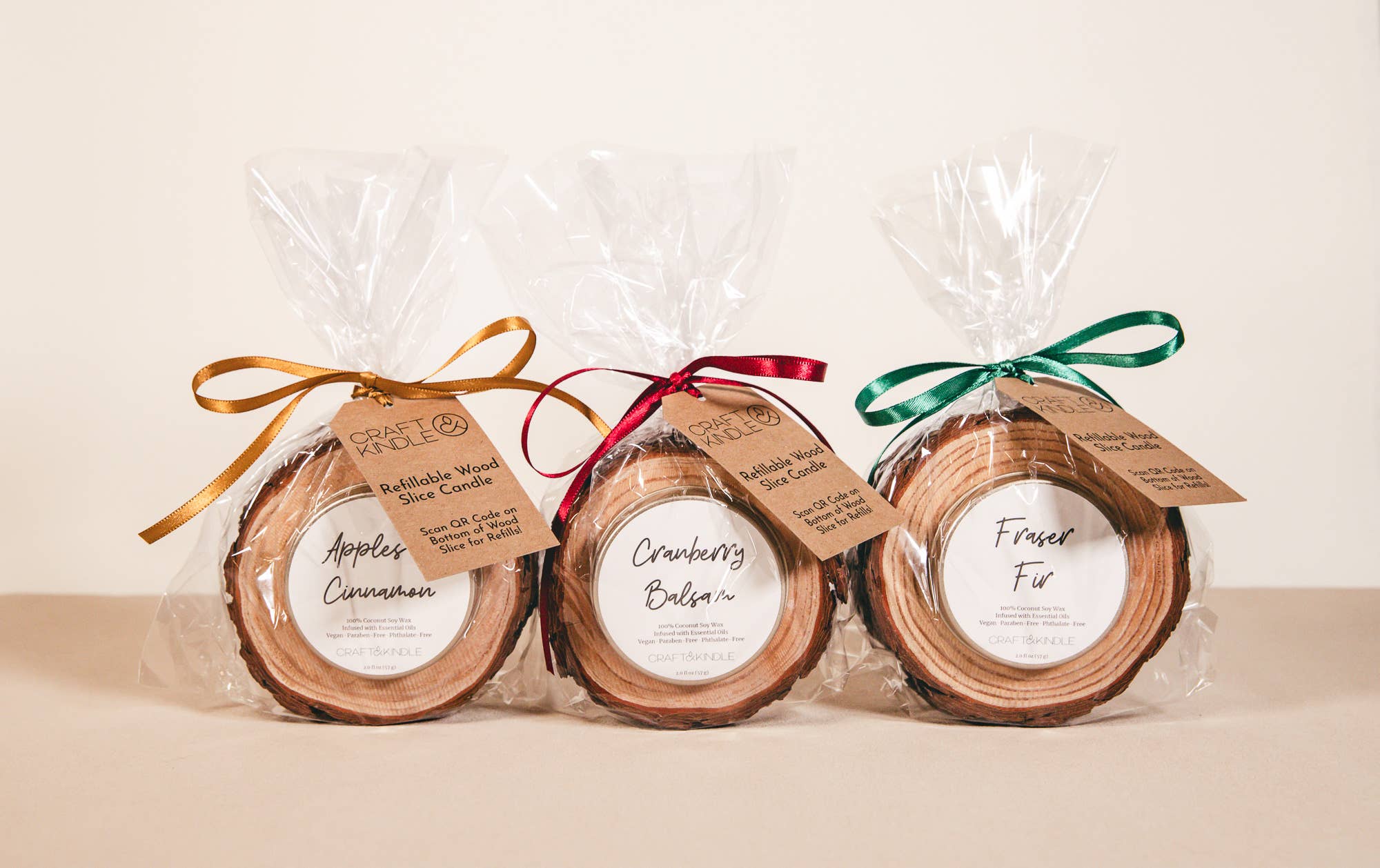 Three wooden candle slices in clear cellophane bags with colorful ribbons and labels on a beige background.
