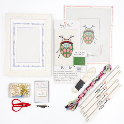 The Beetle kit components on a white background. There is a white frame with a mat with a decorative blue watercolor bamboo border, the canvas with the design, an instructional brochure, colorful threads, tacks, red scissors, a Poppy Monk needle minder, a needle threader, and four Poppy Monk stretcher bars.