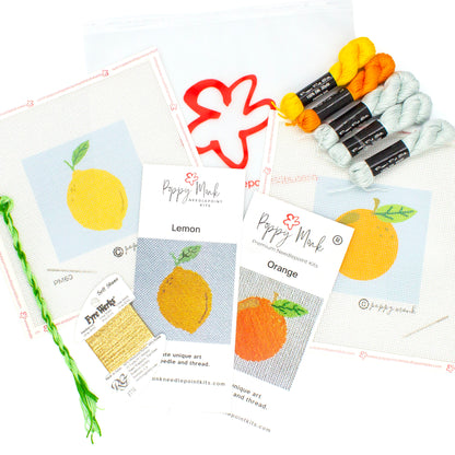 A flat-lay of two needlepoint kits and thread on a white background. Visible items include a canvas with a lemon printed on it, a canvas with an orange printed on it, printed brochure instructions for both, and threads laying on top of a clear Poppy Monk bag with the poppy logo on it.