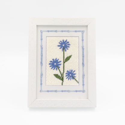 The Cornflower kit stitched and in a white frame with a mat with a decorative blue watercolor bamboo border.