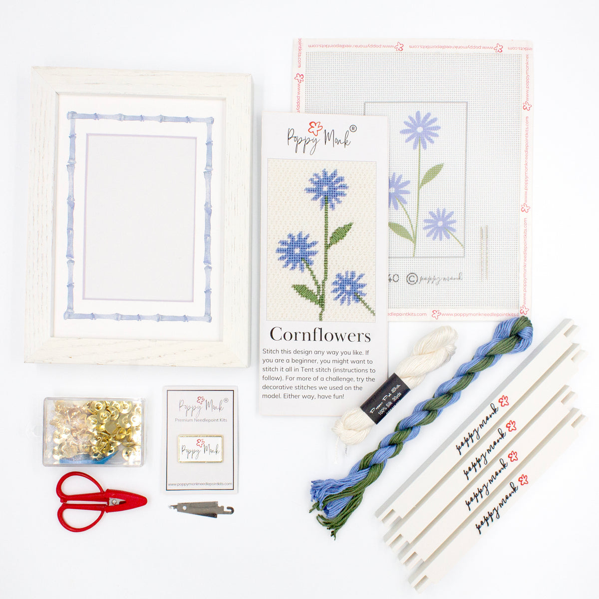 The Cornflower kit components on a white background. There is a white frame with a mat with a decorative blue watercolor bamboo border, the canvas with the design, an instructional brochure, colorful threads, tacks, red scissors, a Poppy Monk needle minder, a needle threader, and four Poppy Monk stretcher bars.