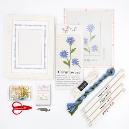 The Cornflower kit components on a white background. There is a white frame with a mat with a decorative blue watercolor bamboo border, the canvas with the design, an instructional brochure, colorful threads, tacks, red scissors, a Poppy Monk needle minder, a needle threader, and four Poppy Monk stretcher bars.