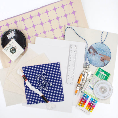 The Finishing Kit components on a white background. This includes blue and white gingham fabric and matching cording, four squares of batting, white thread, cardstock, a ruler, a compass, sewing clips and pins, fishing line, needle, needle threader, instruction booklet, and canvas carry bag.