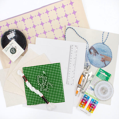 The Finishing Kit components on a white background. This includes green and white gingham fabric and matching cording, four squares of batting, white thread, cardstock, a ruler, a compass, sewing clips and pins, fishing line, needle, needle threader, instruction booklet, and canvas carry bag.