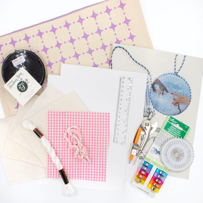 The Finishing Kit components on a white background. This includes pink and white gingham fabric and matching cording, four squares of batting, white thread, cardstock, a ruler, a compass, sewing clips and pins, fishing line, needle, needle threader, instruction booklet, and canvas carry bag.