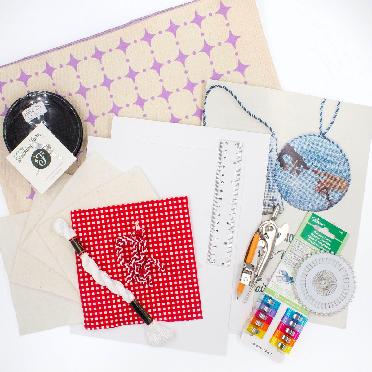 The Finishing Kit components on a white background. This includes red and white gingham fabric and matching cording, four squares of batting, white thread, cardstock, a ruler, a compass, sewing clips and pins, fishing line, needle, needle threader, instruction booklet, and canvas carry bag.