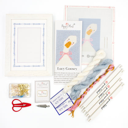 The Lucy Goosey kit components on a white background. There is a white frame with a mat with a decorative blue watercolor bamboo border, the canvas with the design, an instructional brochure, colorful threads, tacks, red scissors, a Poppy Monk needle minder, a needle threader, and four Poppy Monk stretcher bars.