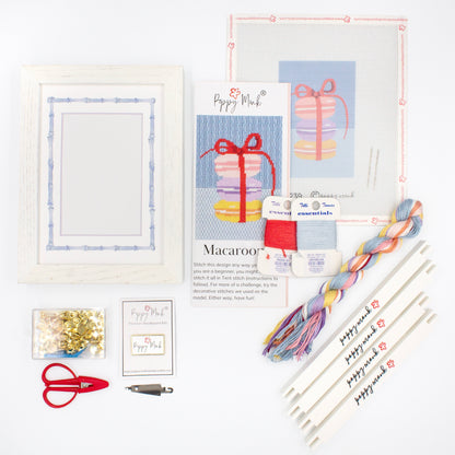 The Macaroons kit components on a white background. There is a white frame with a mat with a decorative blue watercolor bamboo border, the canvas with the design, an instructional brochure, colorful threads, tacks, red scissors, a Poppy Monk needle minder, a needle threader, and four Poppy Monk stretcher bars.