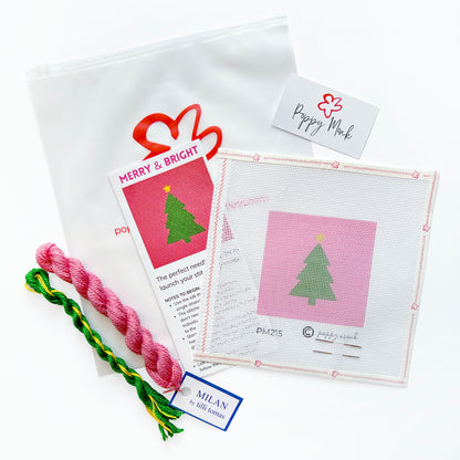 A Christmas-themed needlepoint kit featuring a white mesh canvas with a pink background and green tree design, a pink and green braided cord, a white drawstring pouch, and printed instruction cards on a white background.