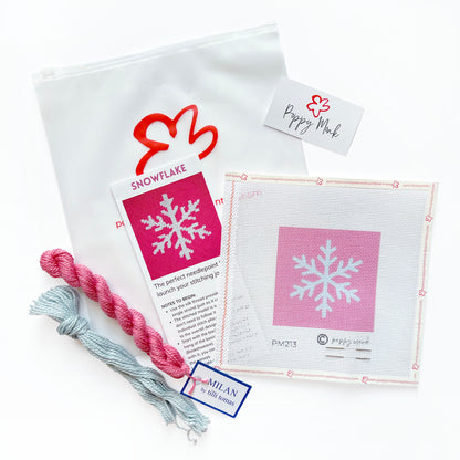 A craft kit laid out on a white background, including a white canvas bag with a red logo, a printed instruction card titled SNOWFLAKE, a pink-and-white swatch card, two skeins of thread in pink and light blue.