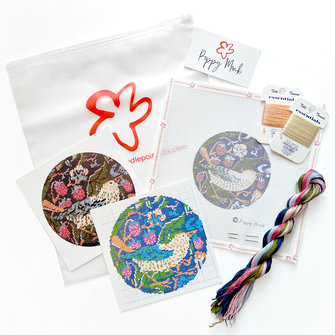 A needlepoint kit laid out on a white background, including patterned fabric discs, thread spools, a braided multicolor thread bundle, a white pouch with a red outline logo.