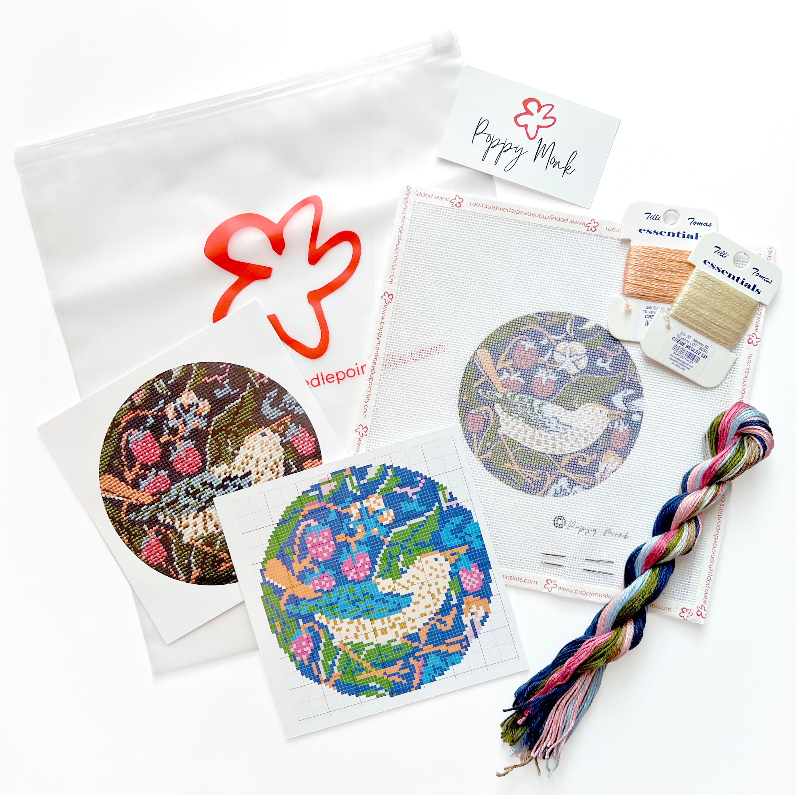 A needlepoint kit laid out on a white background, including patterned fabric discs, thread spools, a braided multicolor thread bundle, a white pouch with a red outline logo.