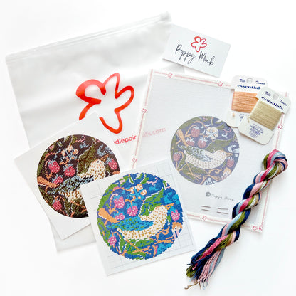 A needlepoint kit laid out on a white background, including patterned fabric discs, thread spools, a braided multicolor thread bundle, a white pouch with a red outline logo.