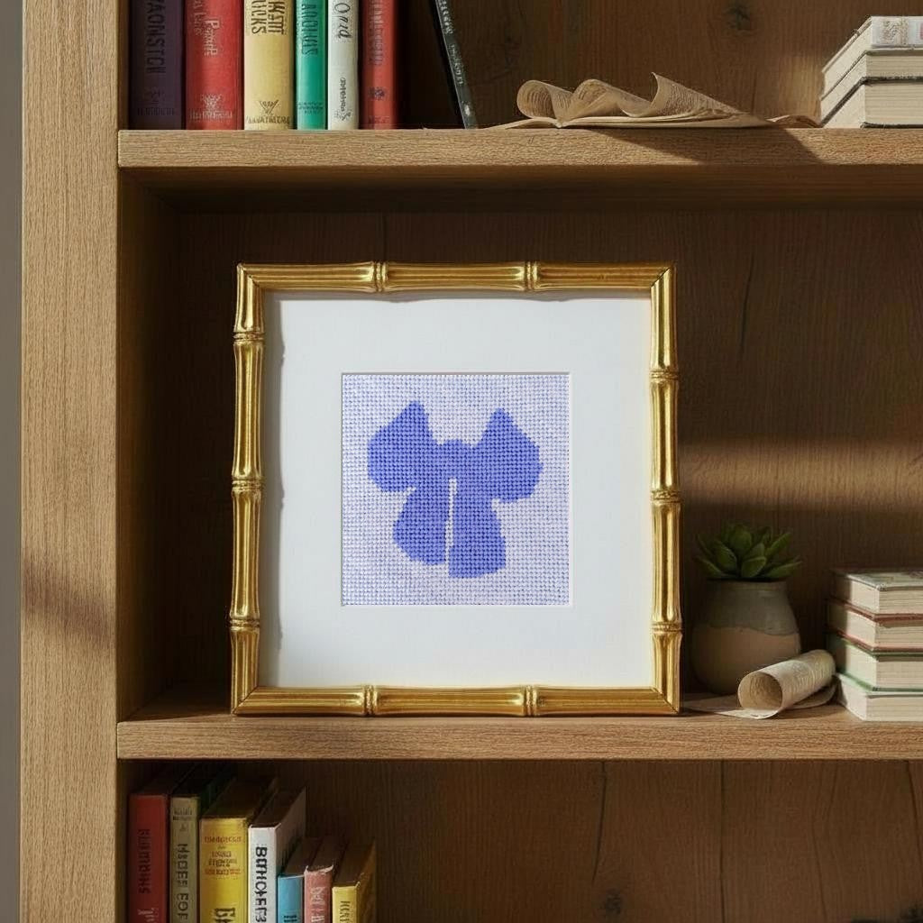 Framed needlepoint with a blue bow design on a wooden shelf with books and a plant.