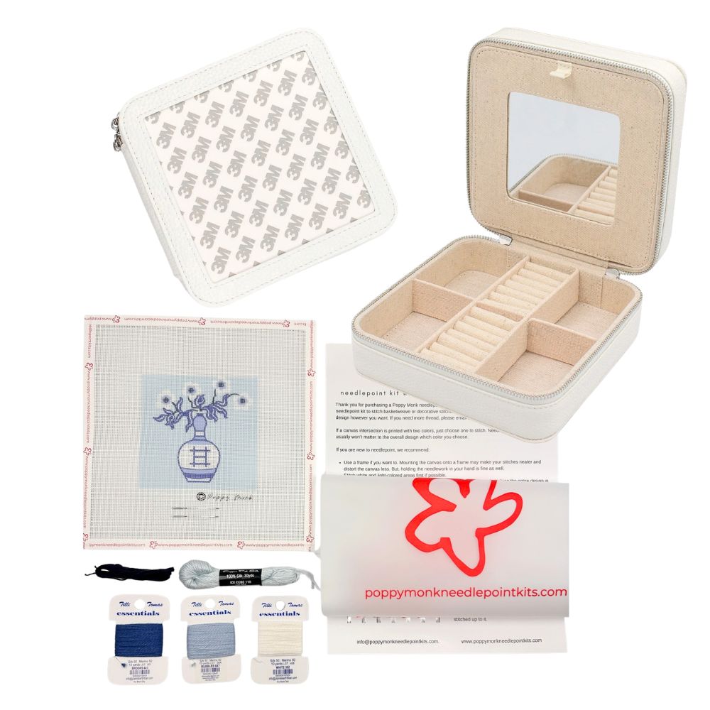 A needlepoint kit set displayed on a white background, including an open cream-colored storage case with a mirror, a folded pattern sheet, printed instructions, three fabric squares in blue, gray, and white, a black marker, gray thread, and a branded card reading poppymonkneedpointkits.com.