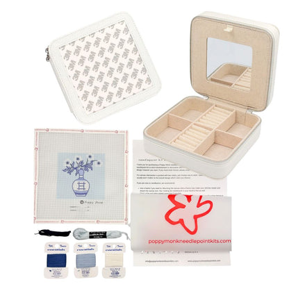 A needlepoint kit set displayed on a white background, including an open cream-colored storage case with a mirror, a folded pattern sheet, printed instructions, three fabric squares in blue, gray, and white, a black marker, gray thread, and a branded card reading poppymonkneedpointkits.com.