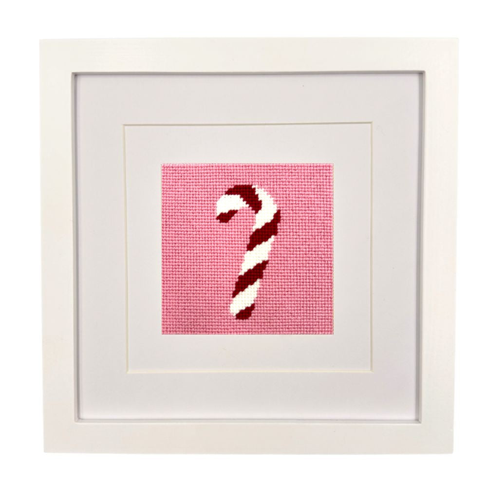 A framed needlepoint artwork featuring a red and white striped candy cane on a pink grid background, presented in a white square frame with wide white matting.