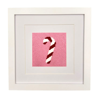 A framed needlepoint artwork featuring a red and white striped candy cane on a pink grid background, presented in a white square frame with wide white matting.