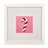 A framed needlepoint artwork featuring a red and white striped candy cane on a pink grid background, presented in a white square frame with wide white matting.