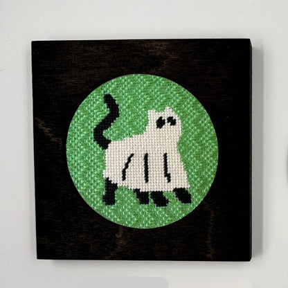 A needlepoint ornament of a cat dressed up as a ghost, displayed in a black frame