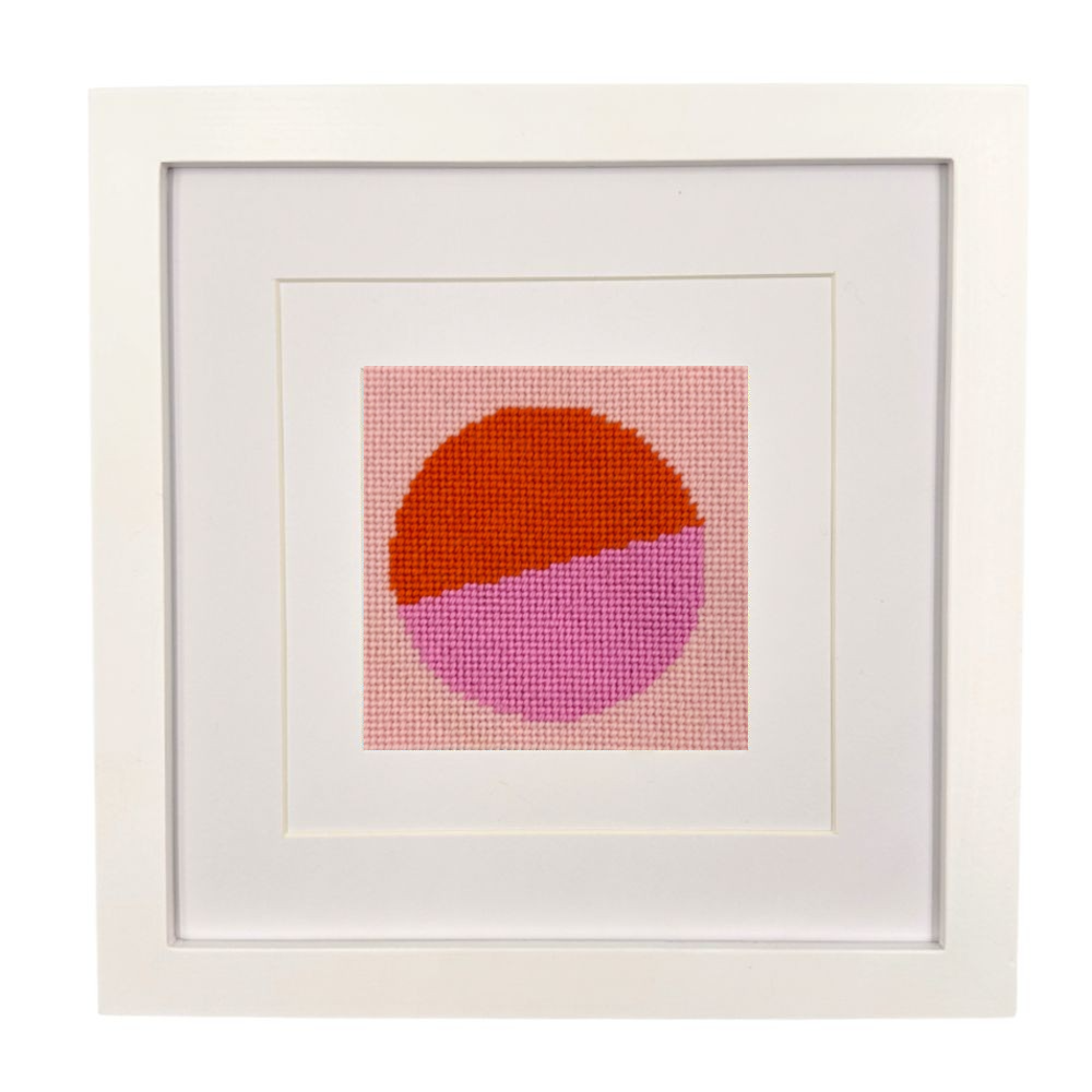 Framed needlepoint with a geometric design of two intersecting circles, one orange and one pink, on a white background.