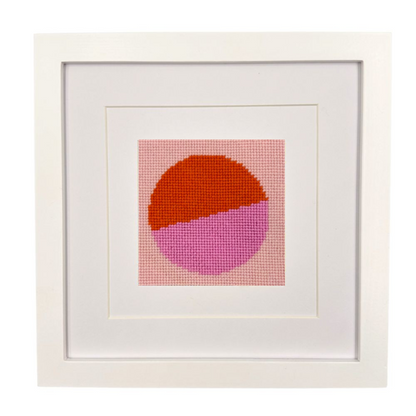 Framed needlepoint with a geometric design of two intersecting circles, one orange and one pink, on a white background.