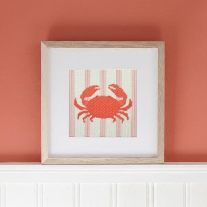 Framed needlepoint crab design on a striped background against an orange wall.