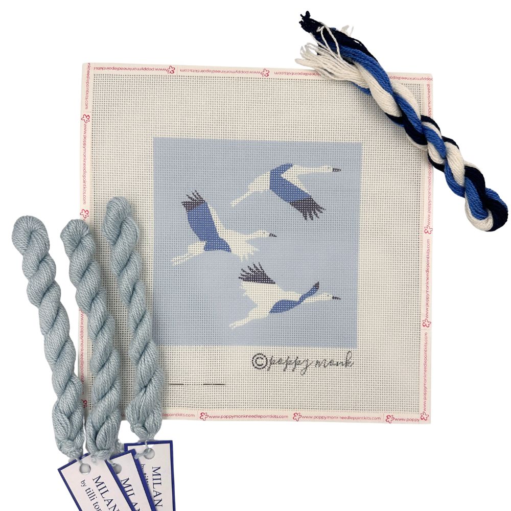 A needlepoint canvas featuring three flying cranes in blue and white on a light blue background, surrounded by a pink stitched border. Three braided threads in light blue, navy, and white are arranged around the canvas, with two tags reading &
