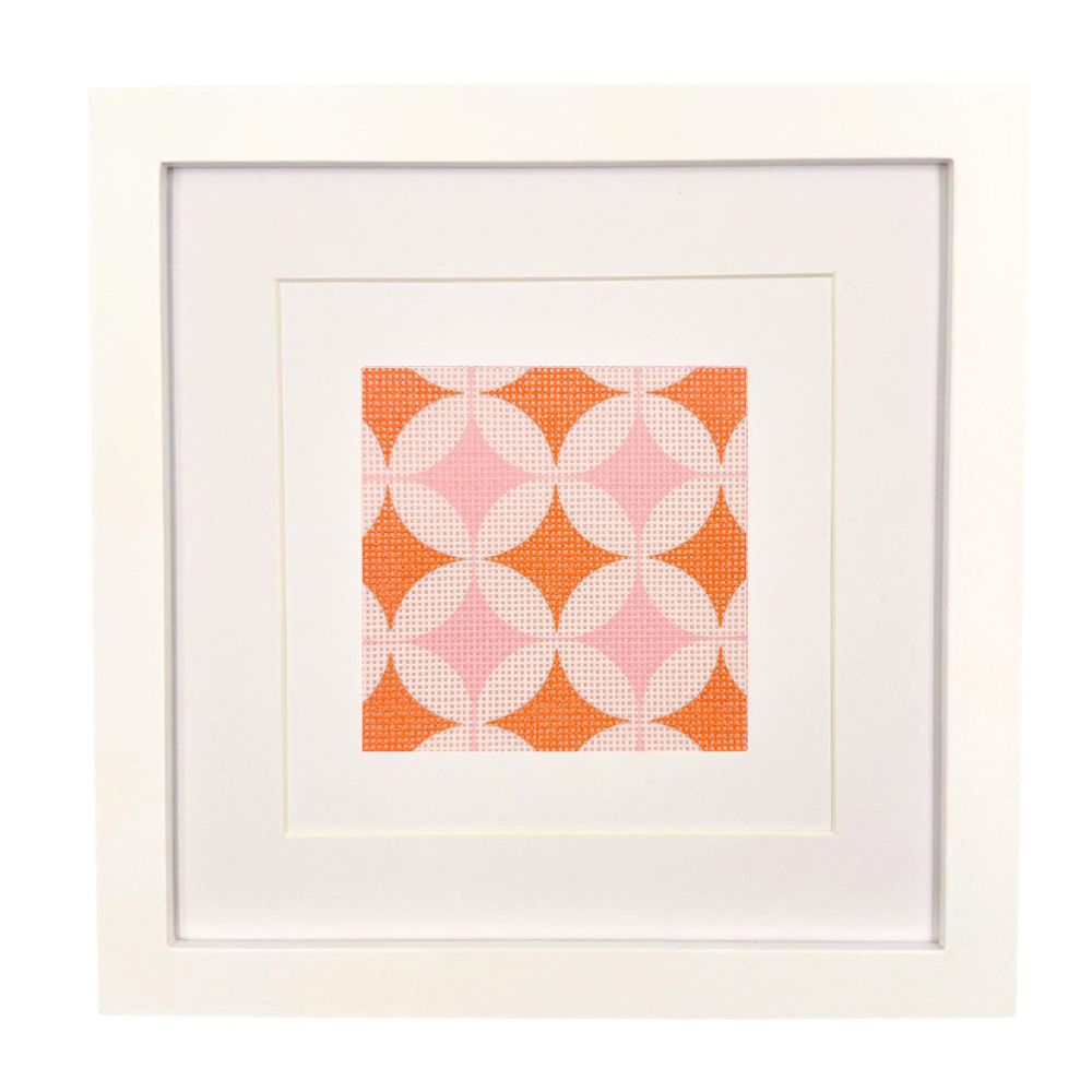 A geometric needlepoint design in orange and pinks shown in a white frame