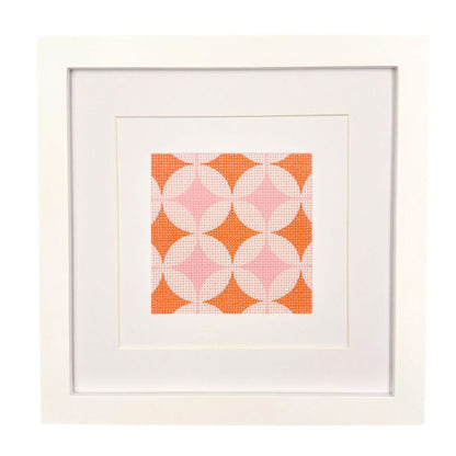 A geometric needlepoint design in orange and pinks shown in a white frame