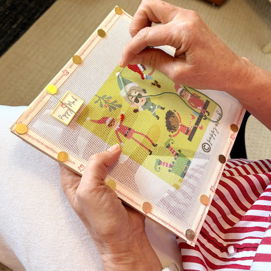 A close-up of hands holding a framed needlepoint canvas with a colorful cartoon design. The person is wearing a red and white striped shirt, and the background shows a neutral indoor setting.
