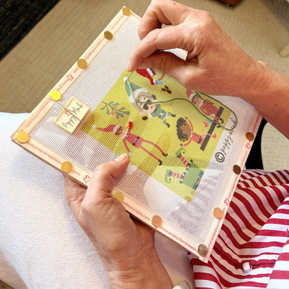 A close-up of hands holding a framed needlepoint canvas with a colorful cartoon design. The person is wearing a red and white striped shirt, and the background shows a neutral indoor setting.