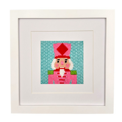 A framed needlepoint canvas featuring a nutcracker character on a teal background, presented in a white square frame with a white mat.