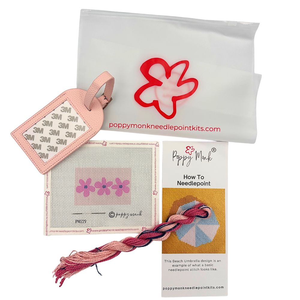 Poppy Monk needlepoint kit components on a white background, including a white canvas pouch with a red flower logo, a pink stitched sample tag, a finished pink and red flower needlepoint canvas, a card explaining needlepoint, and a pink and red braided cord.