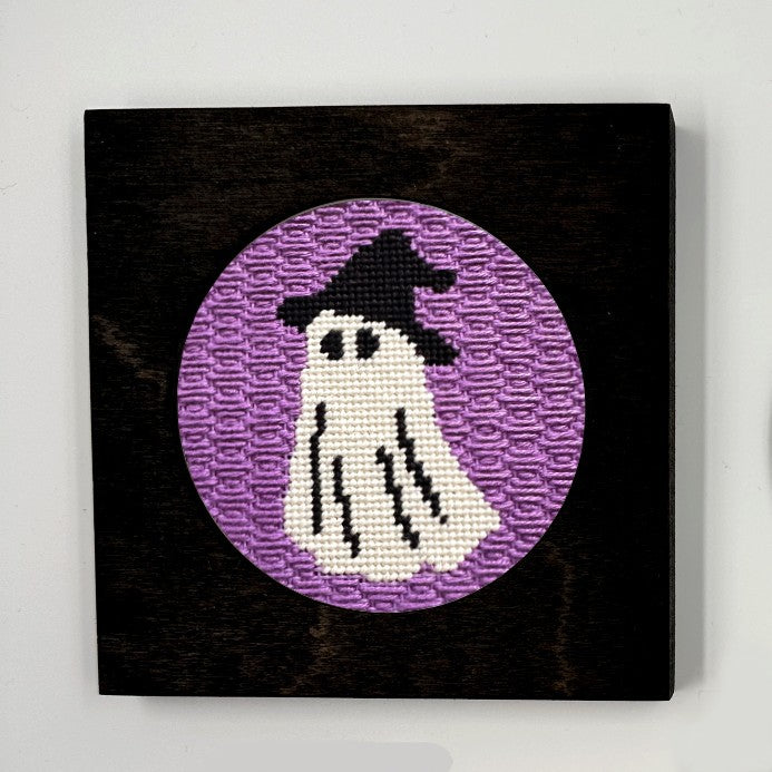 A halloween needlepoint round of a ghost dressed up as a witch, displayed in a frame