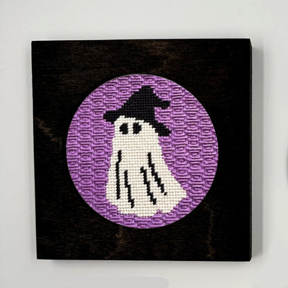 A halloween needlepoint round of a ghost dressed up as a witch, displayed in a frame