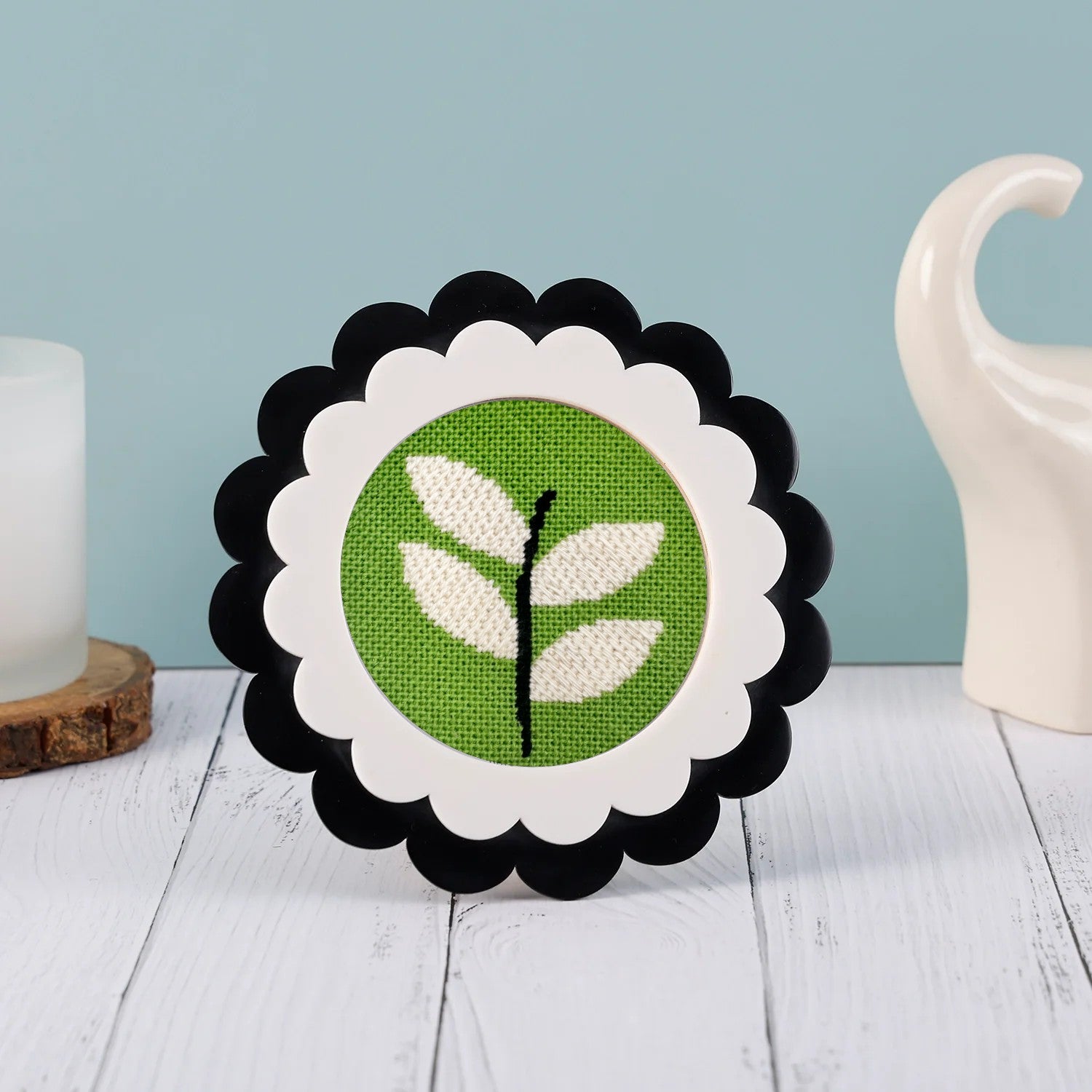 A green and white round needlepoint design displayed in a scalloped frame