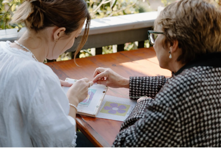 Two women sitting at a table outdoors, one showing something to the other.