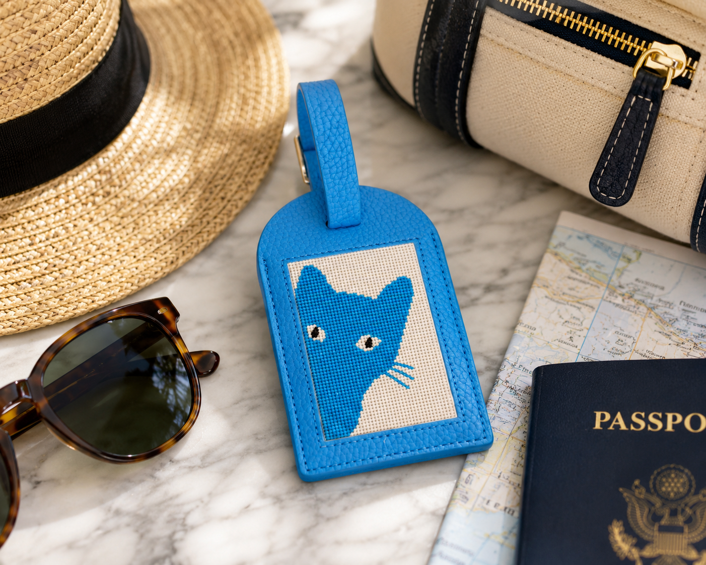 A blue needlepoint luggage tag with cat design shown with travel items
