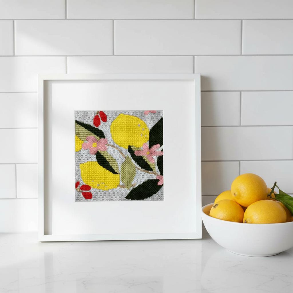 A frsamed lemon needlepoint design shown on a kitchen counter next to a bowl of lemons