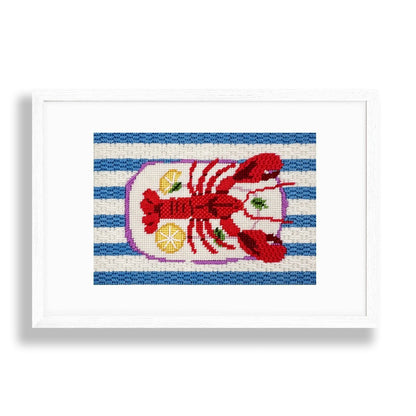 A framed needlepoint featuring a red lobster on a blue and white striped background, enclosed in a white frame against a white wall.