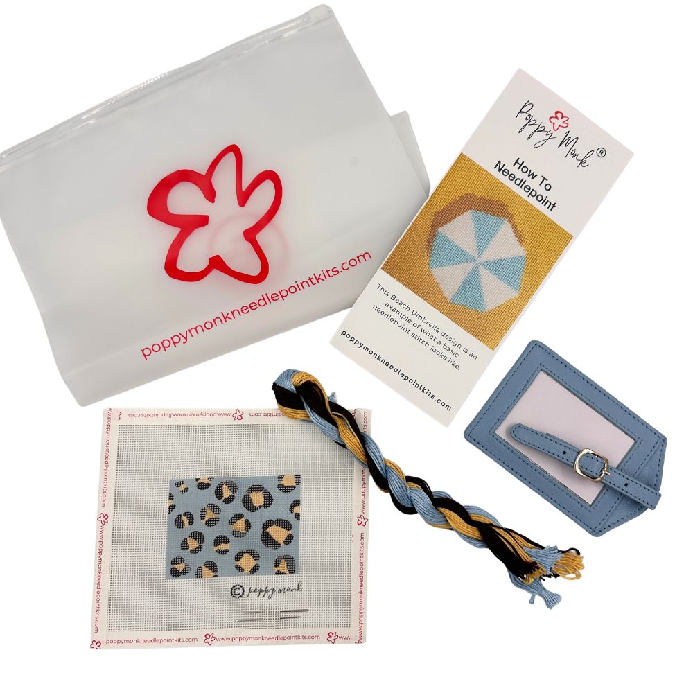 A Poppy Monk needlepoint kit laid out on a white background, including a translucent white pouch with a red flower outline and website print, a printed how-to card, a blue leatherette strap with a metal ring, a square canvas with a leopard-print sample, and a braided cord in blue, tan, black, and white.