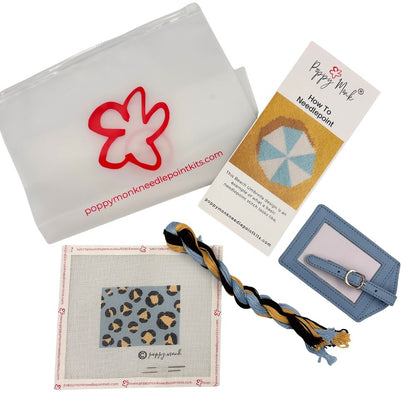 A Poppy Monk needlepoint kit laid out on a white background, including a translucent white pouch with a red flower outline and website print, a printed how-to card, a blue leatherette strap with a metal ring, a square canvas with a leopard-print sample, and a braided cord in blue, tan, black, and white.