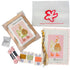 Poppy Monk Needlepoint Kits duck and duck needlepoint kit components laid out on a white background, including a framed finished piece, a white plastic bag with the red Poppy Monk logo and website, two pink skeins of embroidery floss, four small fabric squares, and a canvas with a duck design and braided yellow and white thread.
