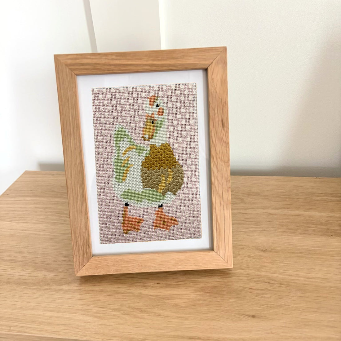 A framed needlepoint artwork of a duck, displayed on a light wood surface against a plain off-white wall. The piece features a light brown duckling with orange beak and feet, a green body, and a white and brown patterned background, enclosed in a light oak frame with a white mat.